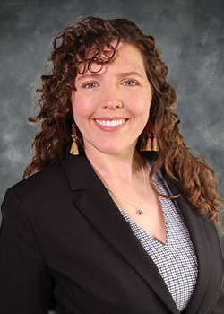 Woman with long, wavy reddish-brown hair wearing a black suit jacket over a black and white patterned blouse