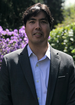 Man with dark brown hair wearing a gray sport coat over blue checkered button up