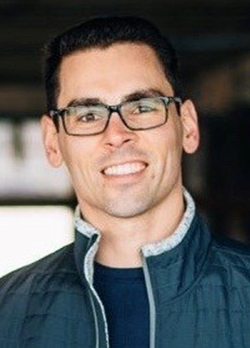 Man with short, cropped hair, wearing rectagular prescription glasses with a navy vest jacket over navy t-shirt