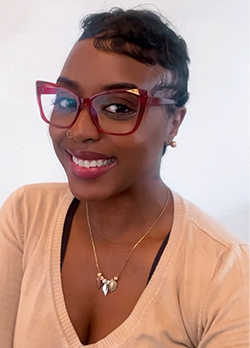 Woman with short, black hair, wearing red rimmed designer oversized glasses and a dark yellow, v-neck sweater